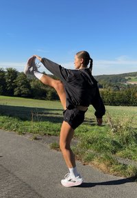 Chubasquero negro, pantalones cortos negros, pierna levantada con calcetas deportivas blancas y rosas, de pie sobre un sendero pavimentado con vegetación de fondo.
