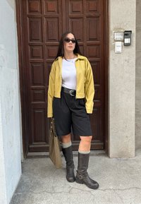 Gul jakke, hvit skjorte, sorte shorts, brune støvler til kneet, og grå kne-sokker, med en beige veske og bakgrunn av en tre dør.
