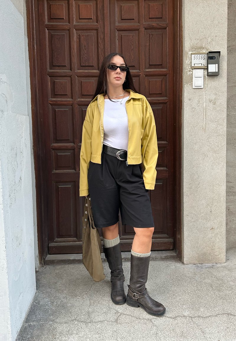 Giacca gialla corta, camicia bianca, pantaloni corti neri, stivaletti marroni al ginocchio e calze grigie fino al ginocchio, con una borsa beige e uno sfondo con porta di legno.
