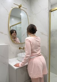 Specchio ovale con cornice dorata sopra un lavabo a ciotola bianca su un piano in marmo chiaro; completo in tessuto rosa liscio con dettagli testurizzati.