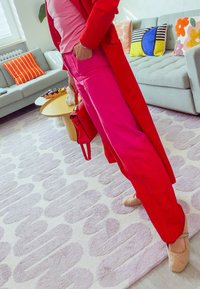 Červený dlouhý kabát, růžová halenka a široké růžové kalhoty. Béžové baleríny, červená kabelka. Světle šedá pohovka s dekorativními polštáři v pozadí.