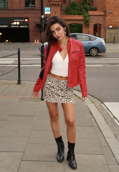 Chaqueta de cuero sintético roja, top corto blanco, falda mini de estampado de leopardo y botas negras hasta el tobillo. De pie en la acera con un fondo urbano.