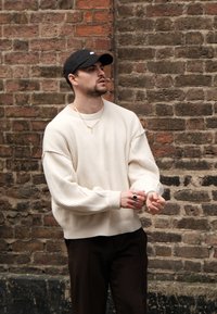 Pulover supradimensionat de culoare crem, cu dungi, și umeri lăsați, asortat cu pantaloni ciudați maro închis. Accesorii incluse: coliere aurii suprapuse și inele.