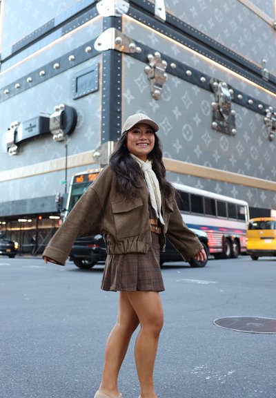 Chaqueta de ante marrón con grandes bolsillos, falda de cuadros marrones con estampado, bufanda clara y gorra beige. De fondo, un gran baúl de Louis Vuitton.