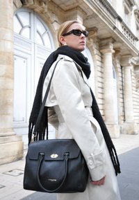 Borsa a tracolla nera in pelle con una forma strutturata, hardware dorato e finitura testurizzata, indossata con un trench beige e una sciarpa nera.