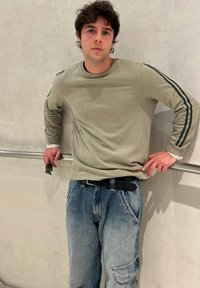 Jeune homme aux cheveux bouclés appuyé sur une rambarde, portant une chemise verte à manches longues rayée et un jean bleu délavé, tenant des lunettes de soleil.