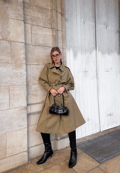 Gabardina caqui con cintura ceñida, botas negras hasta la rodilla y un pequeño bolso negro. La pared presenta una textura de piedra rugosa y puertas de madera.