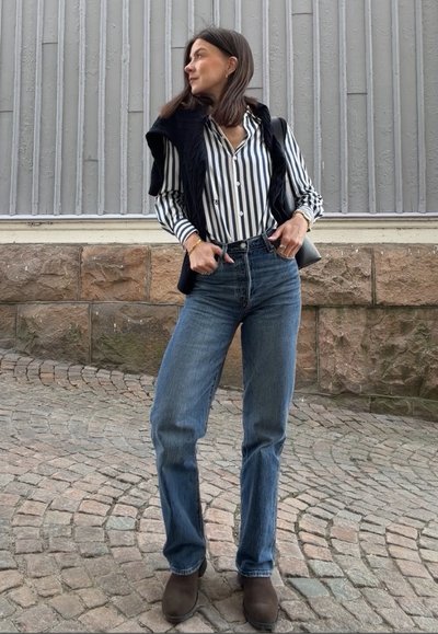 Mujer con camisa de rayas, suéter negro sobre los hombros, vaqueros azules y zapatos marrones, de pie sobre un pavimento empedrado mirando hacia un lado.