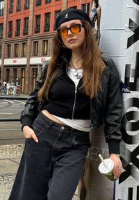 Μαύρο δερμάτινο μπουφάν τύπου bomber πάνω από ένα μαύρο κοντό πουλόβερ, συνδυασμένο με μαύρα φαρδιά τζιν. Τα αξεσουάρ περιλαμβάνουν πορτοκαλί γυαλιά ηλίου και ένα πράσινο ποτό.