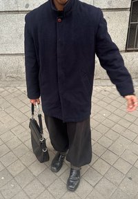 Μαύρο παλτό από μαλλί με όρθιο γιακά, κλείσιμο με μονό κουμπί και πλάγιες τσέπες. Συνδυάζεται με φαρδιά μαύρα παντελόνια και μαύρες δερμάτινες μπότες.