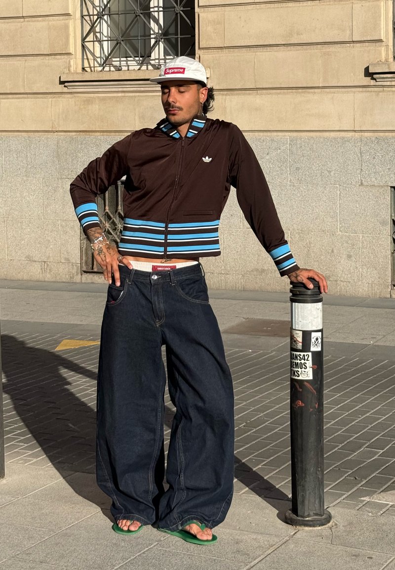Homme portant une casquette blanche, une veste marron avec des rayures bleues, un jean ample foncé et des sandales vertes, appuyé contre un poteau noir sur un trottoir en ville.