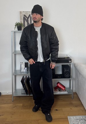 Chaqueta bomber negra, camiseta gris, pantalones vaqueros anchos oscuros y zapatos negros. De pie junto a una estantería con discos de vinilo y altavoces.