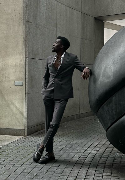 Traje gris de doble botón con una camisa blanca abierta. Zapatos negros con detalle de cadena. Fondo de concreto liso y moderno con una escultura redondeada.