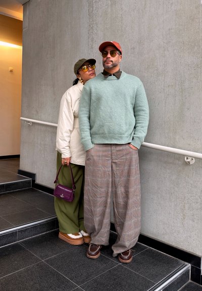 Dos personas con atuendos elegantes de otoño están junto a unas escaleras y una pared de concreto; una lleva un suéter menta y la otra una chaqueta blanca con pantalones verdes.