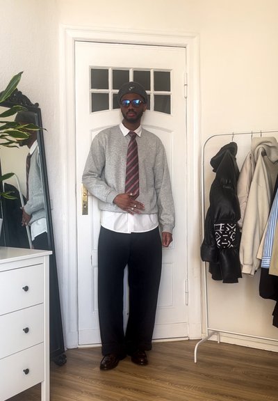 Sudadera gris sobre una camisa blanca con cuello, corbata a rayas roja y negra, pantalones negros de pierna ancha, zapatos marrones y gafas de sol azules.