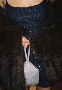Personne portant une robe sombre à paillettes et un manteau en fourrure marron, tenant un petit sac à main argenté étincelant avec une anse.