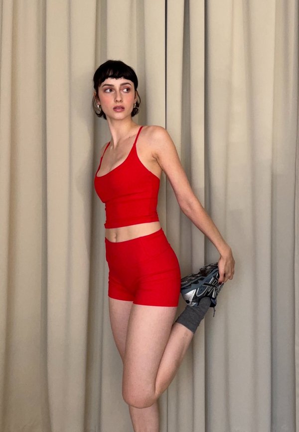 Une femme vêtue d’une tenue de sport rouge étire une jambe vers l’arrière tout en tenant une chaussure de sport, devant des rideaux beiges.