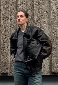 Mustast nahast liibuv helemantel, millel on voltidega krae, kantud üle halli kampsuni. Kujutis hoiab mustast nahast aksessuaari. Tekstuuriga betoonist taust.