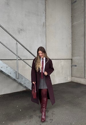 Mujer joven con un abrigo largo oscuro, falda gris, botas burdeos y medias, sosteniendo un bolso a juego, caminando cerca de una pared de hormigón y escaleras de metal.