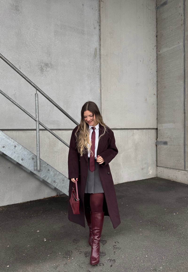 Jeune femme portant un long manteau sombre, une jupe grise, des bottes bordeaux et des collants, tenant un sac à main assorti, marchant près d'un mur en béton et d'un escalier métallique.