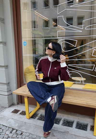 Chaqueta de velour granate con cremallera y rayas blancas, combinada con pantalones vaqueros azules de pierna ancha y zapatillas marrones; sosteniendo una taza de café amarilla.