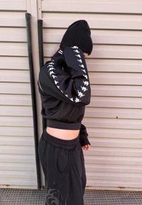 Personne portant un bonnet noir, une veste courte noire avec des logos blancs sur les manches, et un pantalon de jogging noir, debout de profil contre un mur en métal.