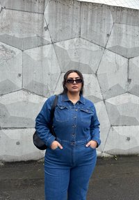 Macacão de ganga azul com fecho frontal de botões, bolsos no peito e mangas longas. Óculos de sol pretos mate e uma bolsa de ombro preta como acessórios.