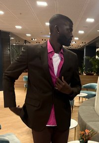 Ruskea bleiseri vaaleanpunaisen paidan ja valkoisen t-paidan päällä, tummien housujen kanssa. Ominaisuuksiin kuuluu tyylikäs muotoilu, näkyvät ompeleet ja minimalistiset metalliosat.