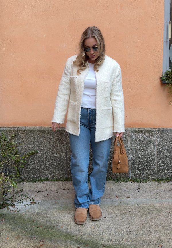Woman wearing sunglasses, white textured jacket, white blouse, blue jeans, tan boots, holding a tan handbag against a peach-coloured wall background.