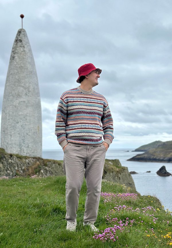 Mann in buntem Pullover und rotem Fischerhut, der auf einer grasbewachsenen Klippe nahe dem Ozean und einem hohen weißen Steindenkmal unter bewölktem Himmel steht.