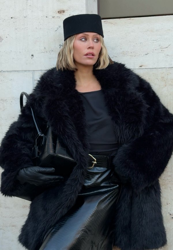 Cappotto in pelliccia sintetica nera con maniche ampie, abbinato a una maglia nera aderente e a una gonna nera lucida con stampa effetto coccodrillo. Gli accessori includono un cappello nero e una borsa.