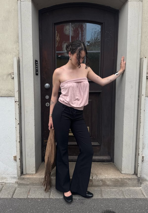 Giovane donna con capelli ricci che indossa un top rosa senza spalline e pantaloni neri, in piedi appoggiata a una porta di legno scuro all'aperto.