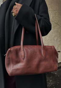 Άτομο με μαύρο παλτό κρατά μια μεγάλη καφέ δερμάτινη τσάντα χειρός από τα λουριά της, με δαχτυλίδια ορατά στα δάχτυλα.