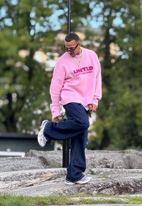 Svijetlo ružičasti pleteni pulover s natpisom "UNTLD", tamnoplave široke traperice i šarene sportske tenisice, stoji na stjenovitom terenu.