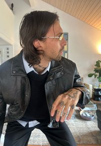 Læderbomberjakke med høj krave, sort farve. Lagdelt over en mørk sweater og en lyseblå skjorte med krave. Bemærkelsesværdige tatoveringer på hænderne.