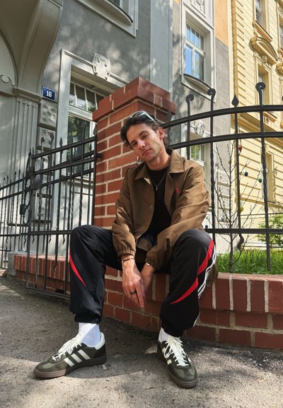 Chaqueta ligera marrón, camisa negra, pantalones deportivos negros con rayas rojas y zapatillas verdes con acentos blancos. Fondo de ladrillo y valla de metal.