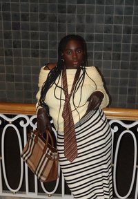 Femme aux longs cheveux tressés portant un pull à épaules dénudées crème, une jupe rayée, une cravate rayée marron, des gants en cuir, et tenant un sac à main en tartan marron.