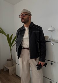 Chaqueta de cuero negra con costuras en beige que contrastan, combinada con una camiseta henley color crema, pantalones beige claros y guantes negros.