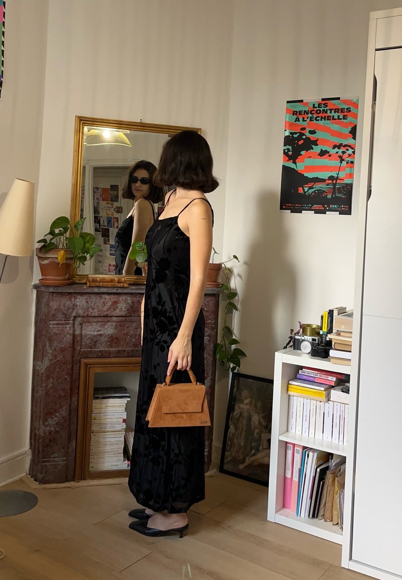 Robe en velours noir avec motif floral, sac à main en daim tan, chaussures noires. Le reflet dans le miroir montre des détails supplémentaires et la décoration intérieure.