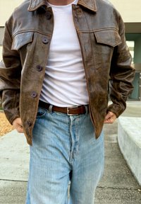 Veste en cuir marron avec fermetures à boutons, deux poches avant et coutures contrastées, portée avec un t-shirt blanc côtelé et un pantalon en denim bleu.