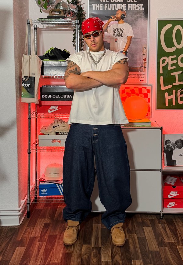 Mann mit Tattoos, der ein rotes Bandana, Sonnenbrille, weisses ärmelloses Shirt, weite Jeans und Stiefel trägt, steht mit verschränkten Armen in einem Raum mit Sneakers und Postern.