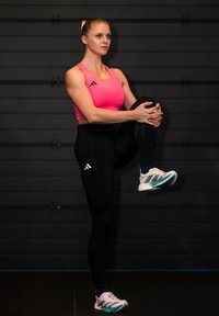 Femme en haut de sport rose et leggings noirs, debout sur une jambe, tenant le genou levé dans une salle de sport avec un fond sombre.