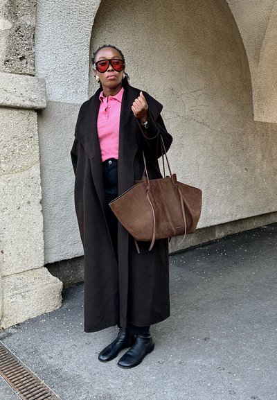 Bolso de ante marrón con asas dobles, abrigo negro de gran tamaño, camiseta de polo rosa, jeans negros y botas negras de tobillo, contra una pared texturizada.