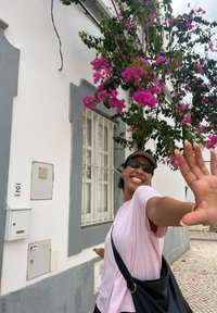 Ružové tričko je oblečené s čiernou taškou, stojí pri bielej stene zdobenej fialovými bougainvilleami. Šedé akcenty na stene.