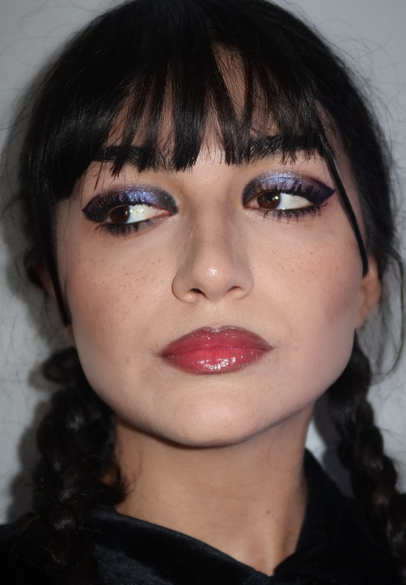 Donna con capelli scuri intrecciati, trucco occhi purpureo glitterato audace, eyeliner a ala, labbra rosse lucide e lentiggini, che guarda a sinistra.