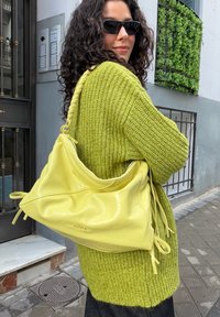 Femme aux cheveux bouclés foncés et lunettes de soleil porte un pull en maille vert et transporte un grand sac à bandoulière en cuir jaune en plein air.