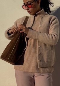 Mulher a usar um suéter e cardigan em malha bege, ajustando uma bolsa de camurça castanha com tachas de latão, contra uma parede clara à luz do sol.