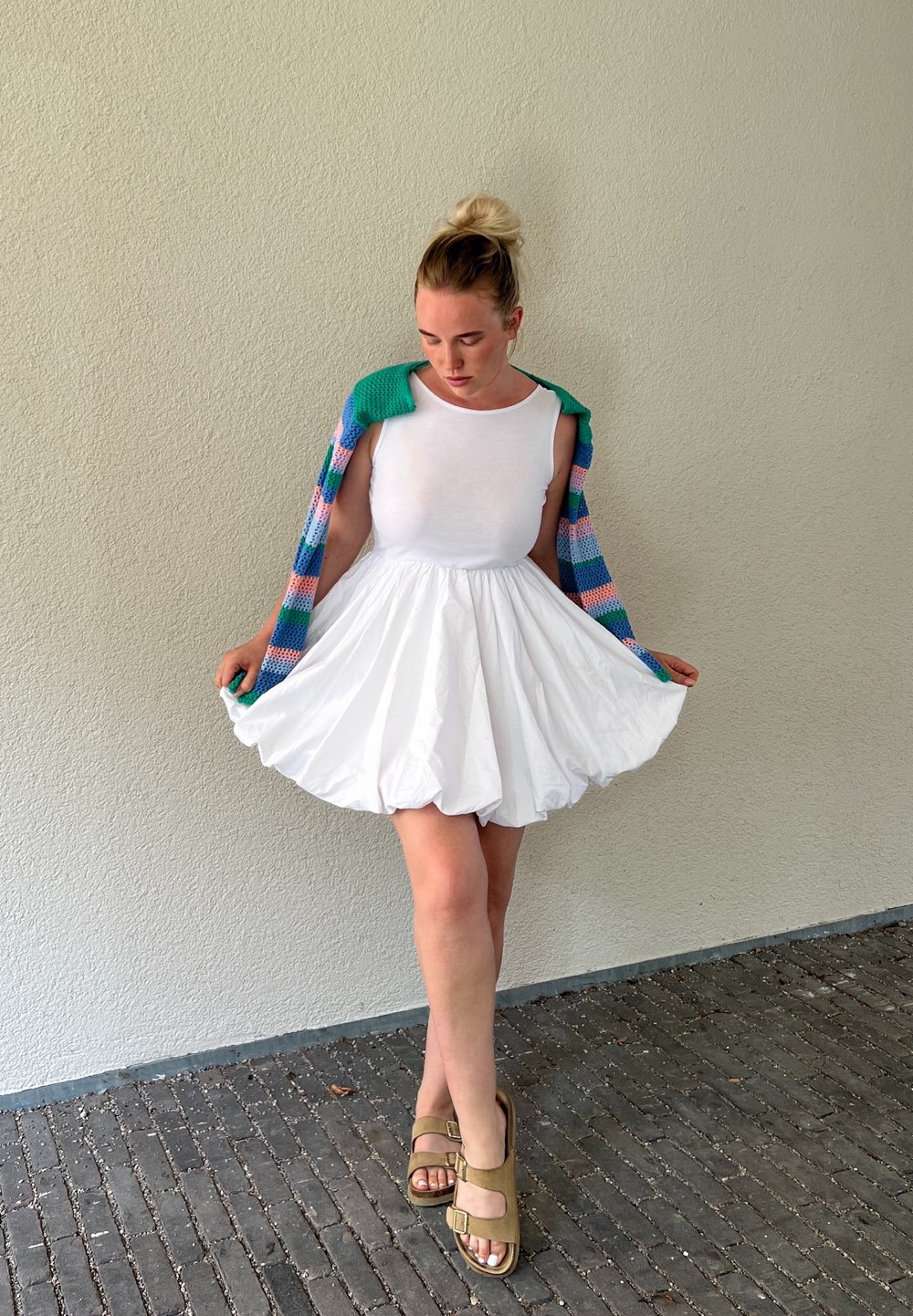 Robe jupe bouffante blanche avec un haut ajusté et un cardigan en tricot coloré drapé sur les épaules. Le mannequin porte des sandales vert olive sur un sol texturé.