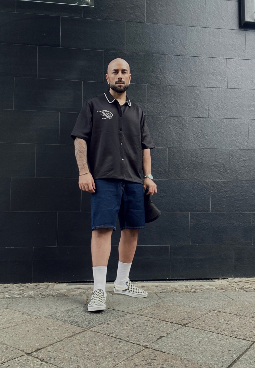Črna kratkoročnega srajca z belim šivanjem, denim kratke hlače, bele nogavice in črno-bele kariraste slip-on čevlje. Enostavna ozadje.