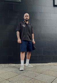 Črna kratkoročnega srajca z belim šivanjem, denim kratke hlače, bele nogavice in črno-bele kariraste slip-on čevlje. Enostavna ozadje.